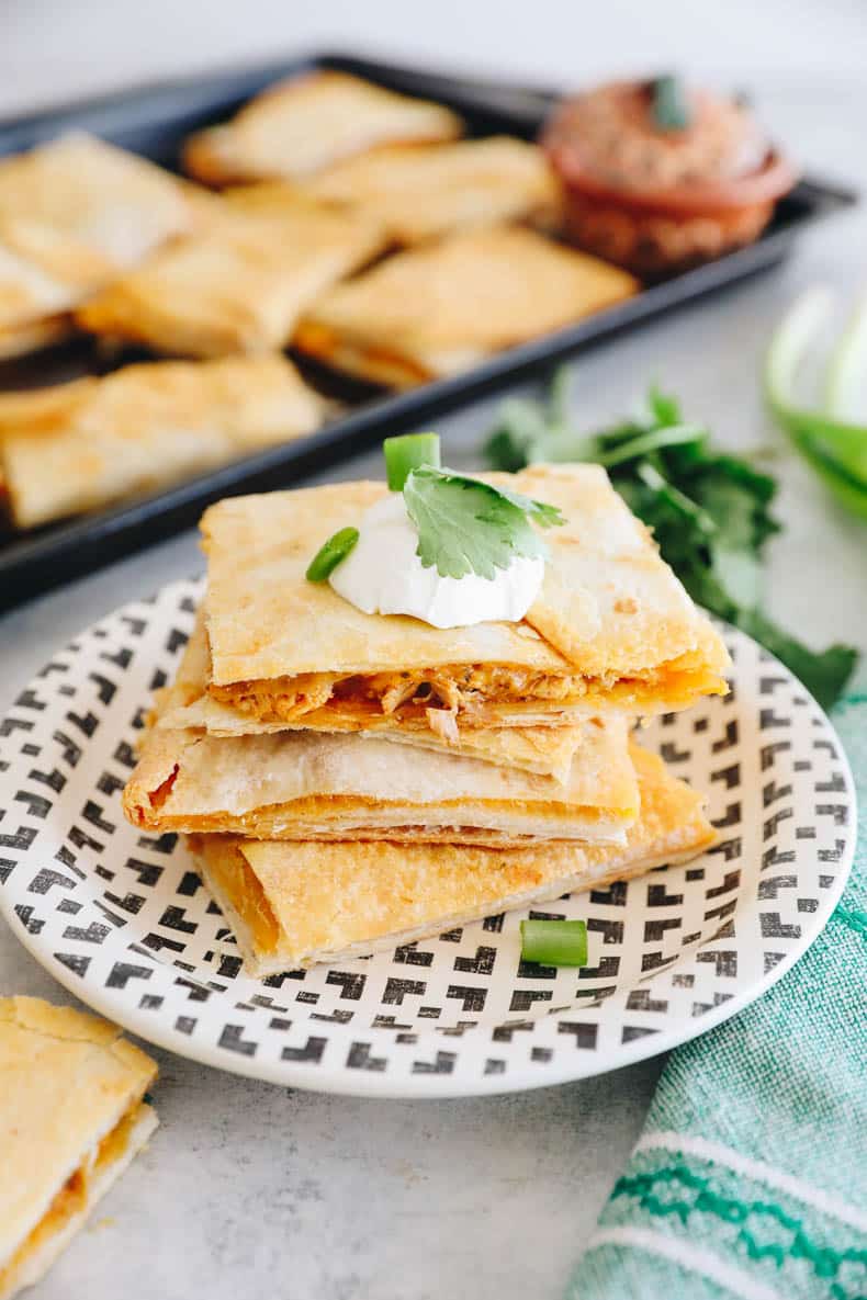 3 quesadillas stacked on top of one another with a sheet pan with baked quesadillas in the background.
